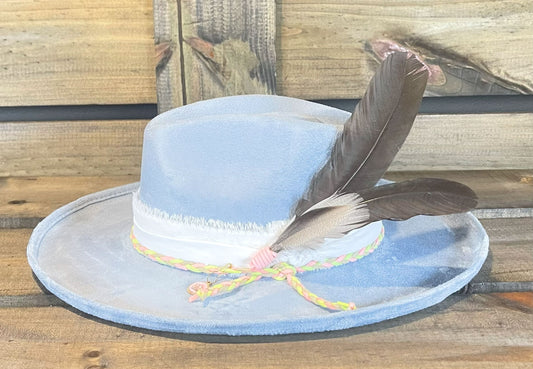 A Carolina Blue vegan suede pencil brim hat wrapped with frayed cotton and wrapped with a braided suede string. Styled with big beautiful turkey feathers and crown charm.